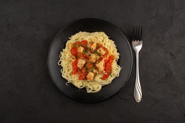 Vista superior de pasta con alitas de pollo y salsa de tomate dentro de la placa negra en el piso oscuro