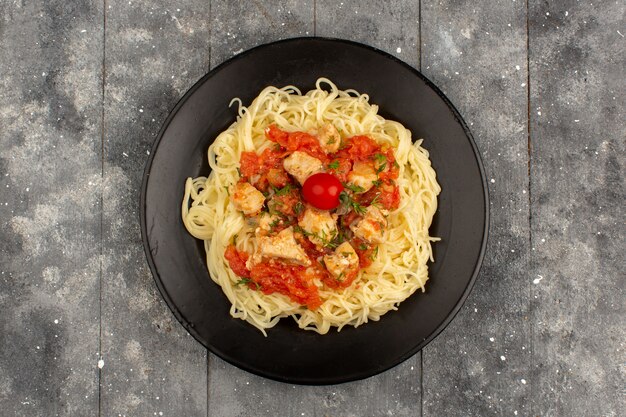 Vista superior pasta cocida con salsas de tomate y alitas de pollo en rodajas dentro de la placa negra en el piso de madera rústico gris