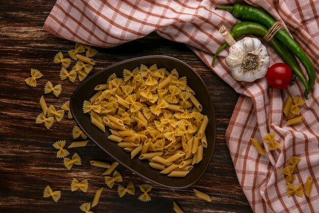 Vista superior de la pasta cruda en un recipiente con ajo, tomate y ají sobre una toalla a cuadros sobre una superficie de madera