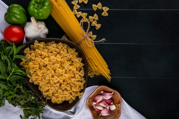 Vista superior de la pasta cruda en un recipiente con espaguetis, tomates, ajo y pimiento con menta sobre una toalla blanca sobre una superficie negra