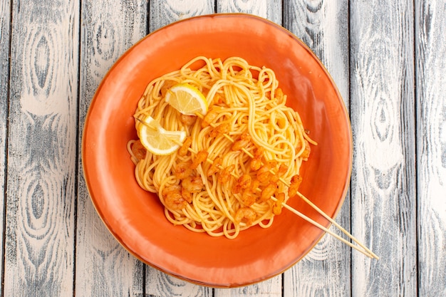 Vista superior de la pasta italiana cocida sabrosa con rodajas de limón y camarones dentro de la placa naranja sobre la superficie rústica de madera gris