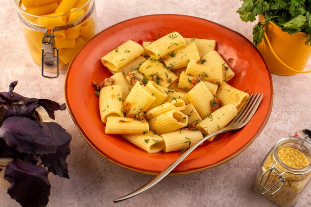 Una vista superior de la pasta italiana cocida sabrosa salada dentro de la placa redonda de naranja con flores y pasta cruda en el escritorio rosa