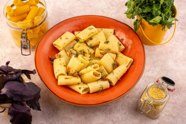 Una vista superior de la pasta italiana cocida sabrosa salada dentro de la placa redonda de naranja con flores y pasta cruda en el escritorio rosa