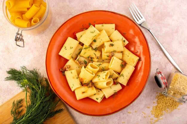 Una vista superior de la pasta italiana cocida sabrosa salada dentro de la placa redonda de naranja con verduras y pasta cruda en el escritorio rosa