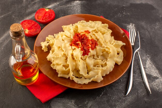 Una vista superior de pasta italiana cocida con salsa de tomate dentro de la placa con cubiertos y tomates en la mesa gris comida pasta italiana