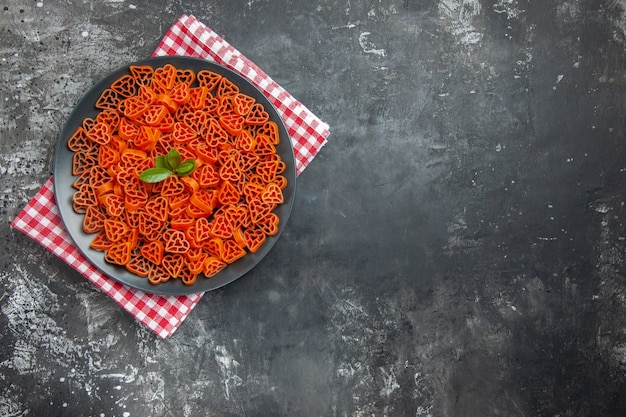 Vista superior de pasta italiana roja en forma de corazón en plato ovalado negro sobre una toalla de cocina en el espacio libre de la mesa oscura