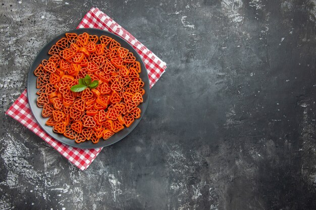 Vista superior de pasta italiana roja en forma de corazón en plato ovalado negro sobre una toalla de cocina en el espacio libre de la mesa oscura