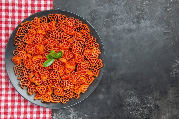 Vista superior de pasta italiana roja en forma de corazón en plato ovalado negro sobre una toalla de cocina en una mesa oscura con espacio libre