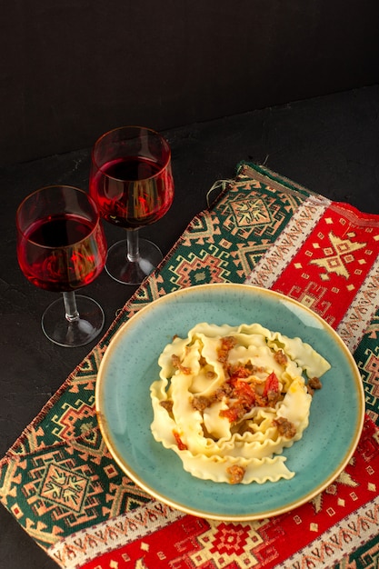 Una vista superior de la pasta de masa cocida sabrosa salada dentro de la placa verde redonda con copas de vino en la alfombra diseñada y escritorio oscuro