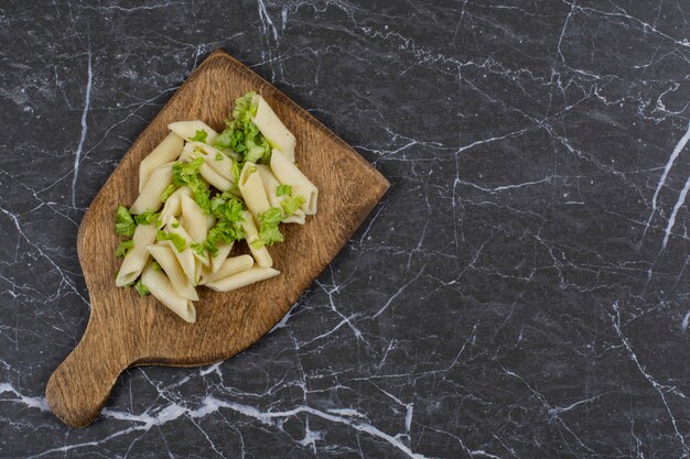 Vista superior de pasta penne con verduras sobre tabla de madera sobre negro.