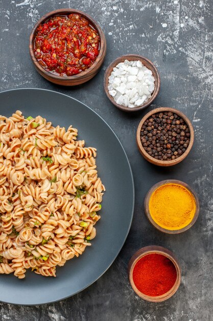 Vista superior de pasta rotini en plato redondo salsa de tomate diferentes especias en tazones pequeños sobre mesa oscura