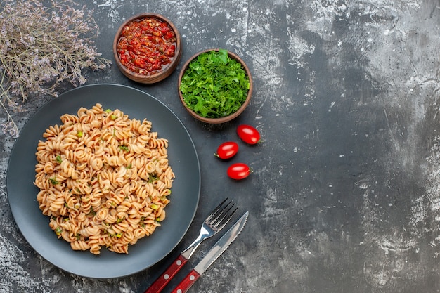 Vista superior de pasta rotini en plato redondo salsa de tomate perejil picado en tazones pequeños tomatotes cherry tenedor y cuchillo en la mesa oscura espacio libre