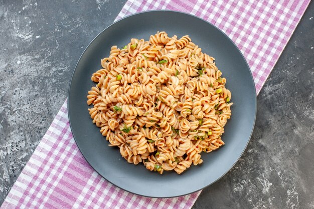 Vista superior de pasta rotini en plato redondo sobre mantel a cuadros blanco rosa sobre superficie oscura