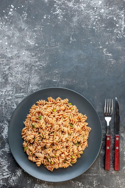 Vista superior de pasta rotini en plato redondo, tenedor y cuchillo en mesa oscura con lugar libre