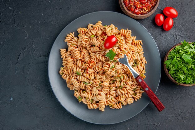 Vista superior de pasta rotini con tenedor de tomate cherry en un plato de perejil y salsa de tomate en tazones sobre una superficie oscura