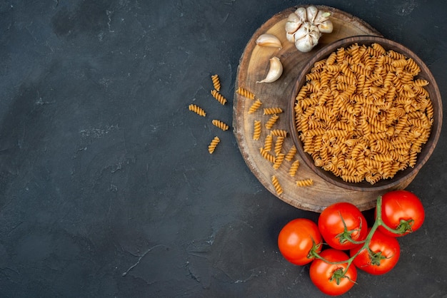 Vista superior de pastas crudas dentro y fuera de tomates de ajo de olla marrón en el lado izquierdo sobre fondo de color azul oscuro