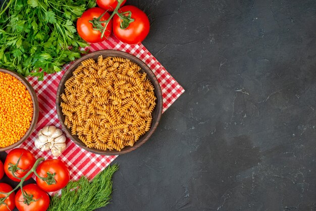 Vista superior de pastas crudas en una tabla de cortar de madera sobre una toalla roja pelada y tomates ajos verdes en el lado derecho sobre fondo oscuro