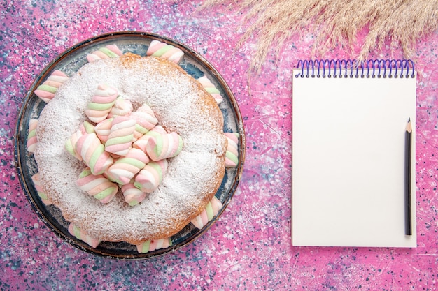 Vista superior de pastel de azúcar en polvo con malvaviscos dulces y bloc de notas en superficie rosa
