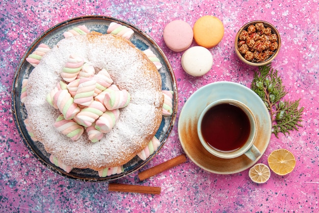 Vista superior del pastel de azúcar en polvo con una taza de té y macarons franceses en la superficie rosa