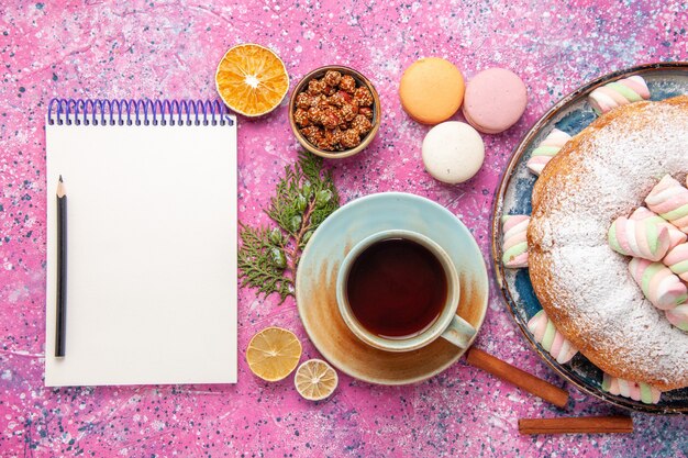 Vista superior del pastel de azúcar en polvo con una taza de té y macarons franceses en la superficie rosa