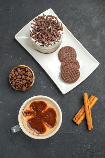 Vista superior de pastel de chocolate y galletas en plato rectangular blanco taza de café tazón de palitos de canela con semillas de café sobre fondo oscuro aislado