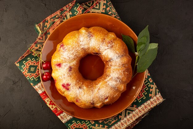 Una vista superior pastel de frutas horneado delicioso redondo con cerezas rojas en el interior y azúcar en polvo dentro de la placa redonda marrón en la oscuridad