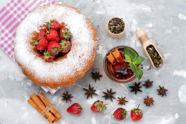 Una vista superior de pastel redondo con azúcar en polvo, fresas rojas, té de canela en el escritorio blanco pastel de frutas de baya