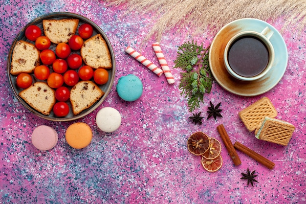 Vista superior de pastel en rodajas con macarons franceses de ciruelas frescas ácidas y una taza de té en el escritorio rosa