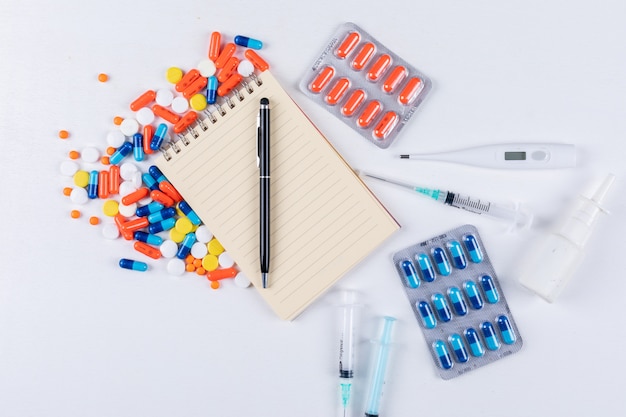 Vista superior de pastillas de colores con bloc de notas, bolígrafo, termómetro, spray nasal y aguja
