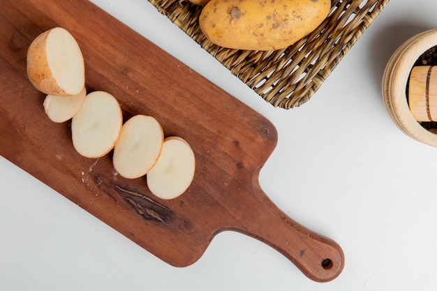 Vista superior de patata cortada y en rodajas en la tabla de cortar con una entera en un plato y pimienta negra sobre una superficie blanca