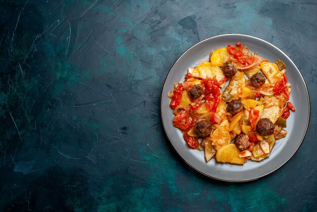 Vista superior de patatas al horno con albóndigas y verduras dentro de la placa sobre el fondo azul oscuro.