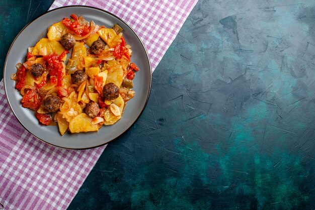 Vista superior de patatas al horno con albóndigas y verduras dentro de la placa sobre el fondo azul oscuro.