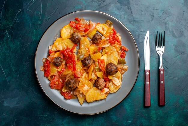 Vista superior de patatas al horno con albóndigas y verduras dentro de la placa sobre el fondo azul oscuro.