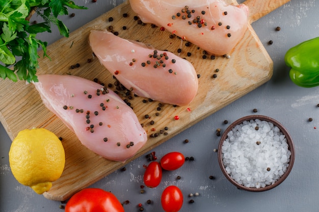 Vista superior pechuga de pollo con pimienta en tabla de cortar con tomates, sal, limón sobre superficie gris