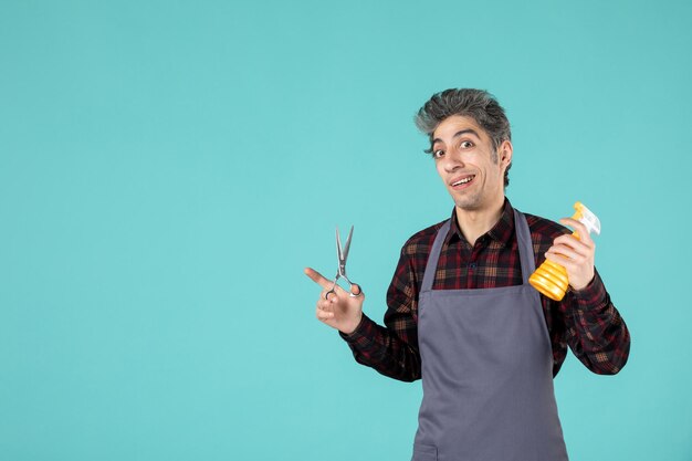 Vista superior del peluquero masculino confundido con delantal gris y sosteniendo una botella de spray de tijeras sobre fondo de color azul suave