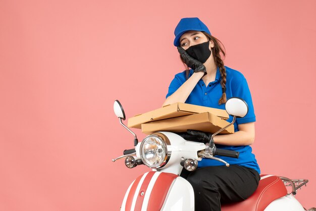Vista superior del pensamiento mensajero femenino con máscara médica y guantes sentado en scooter entregando pedidos en melocotón pastel