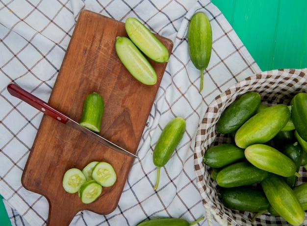 Vista superior de pepino cortado y en rodajas con un cuchillo en la tabla de cortar con los enteros en la cesta sobre tela escocesa y superficie verde