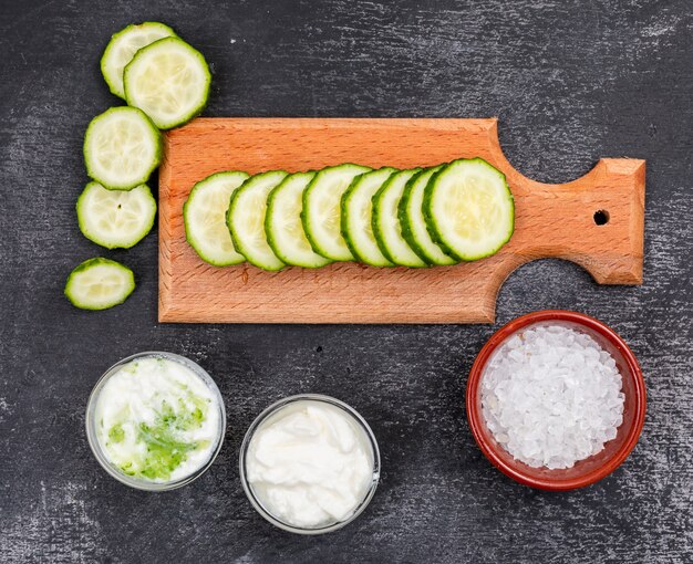 Vista superior de pepino en rodajas con sal y salsas en cuencos pequeños sobre tabla para cortar madera en piedra negra