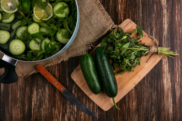 Vista superior de pepinos picados con menta en una sartén con una tabla de cortar y un cuchillo sobre una superficie de madera