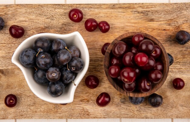 Vista superior de las pequeñas endrinas de fruta amarga azul-negra en un recipiente blanco con cerezas rojas en un recipiente de madera sobre una tabla de cocina de madera sobre un fondo blanco.