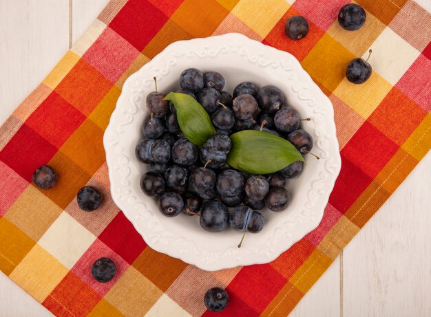 Vista superior de las pequeñas endrinas de fruta amarga azul-negra en un recipiente blanco sobre un mantel a cuadros sobre un fondo blanco.