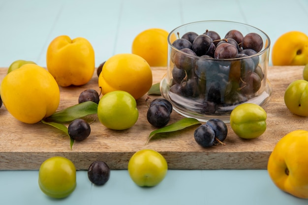 Vista superior de las pequeñas endrinas de fruta amarga azul-negra en un recipiente de vidrio sobre una tabla de cocina de madera con ciruelas cereza verde con melocotones amarillos sobre un fondo azul