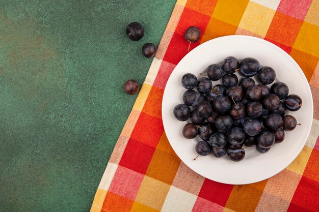 Vista superior de las pequeñas endrinas de fruta amarga de color azul-negro en una placa blanca sobre una tela marcada sobre un fondo verde