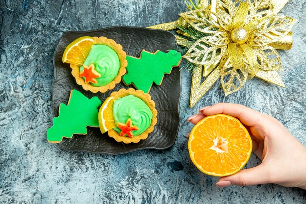 Vista superior pequeñas tartas galletas de árbol de Navidad en placa negra ornamento de Navidad cortado naranja en mano de mujer en mesa gris