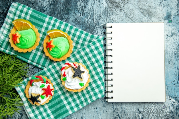 Vista superior de pequeñas tartas de Navidad en el bloc de notas de ramas de pino mantel en mesa gris