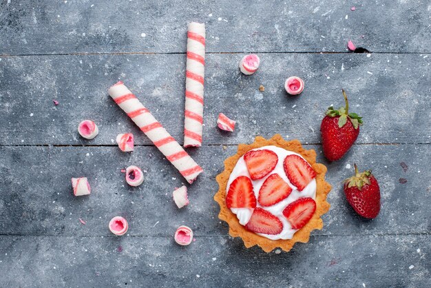 Vista superior de un pequeño pastel cremoso con fresas frescas en rodajas junto con caramelos en el fondo gris pastel de frutas y bayas foto de color dulce hornear