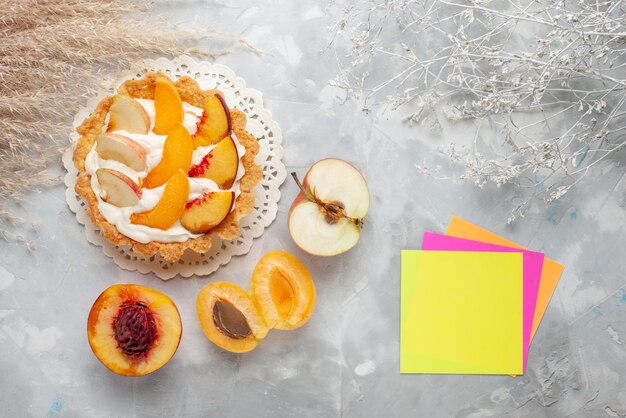 Vista superior del pequeño pastel cremoso con frutas en rodajas y crema blanca junto con albaricoques frescos y melocotones en la galleta de pastel de frutas de escritorio de luz blanca