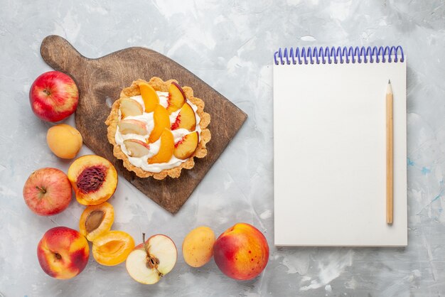 Vista superior del pequeño pastel cremoso con frutas en rodajas y crema blanca junto con el bloc de notas de frutas frescas en el escritorio blanco claro Pastel de frutas Galleta de crema dulce