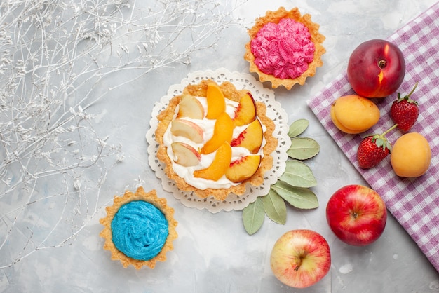 Vista superior del pequeño pastel cremoso con frutas en rodajas y crema blanca junto con pasteles cremosos y frutas en un escritorio ligero, pastel de frutas, galleta, galleta dulce