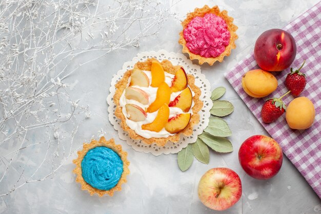 Vista superior del pequeño pastel cremoso con frutas en rodajas y crema blanca junto con pasteles cremosos y frutas en un escritorio ligero, pastel de frutas, galleta, galleta dulce